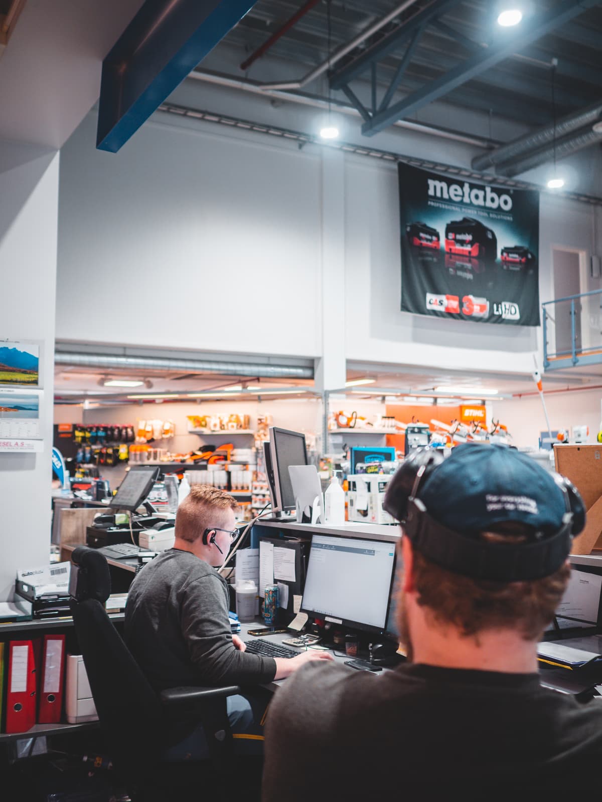 To menn sitter foran pcskjermer bak skranken i butikken.